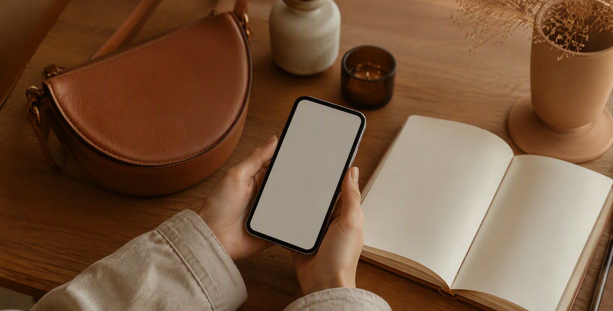 Руки со смартфоном с чистым экраном над деревянным столом, рядом мягкая коричневая сумка-полумесяц и открытый блокнот