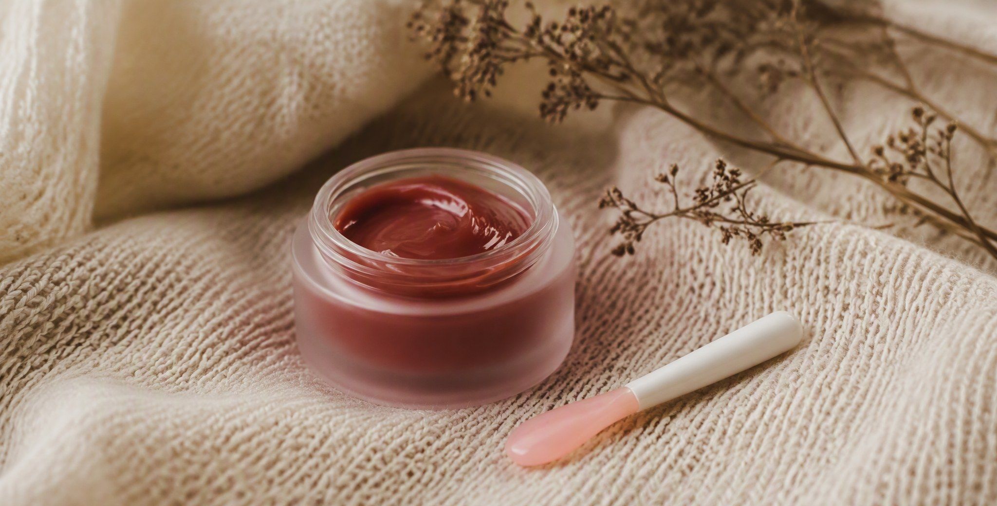 Баночка ночной маски для губ с ягодным оттенком и маленькая лопаточка для нанесения лежат на вязаной ткани